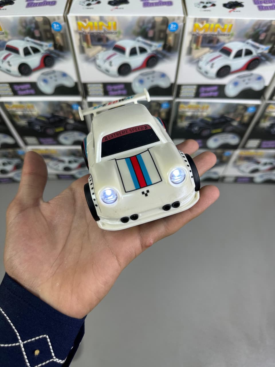 A hand holding a small white toy race car with blue headlights and red, white, and blue racing stripes, with toy boxes stacked in the background.
