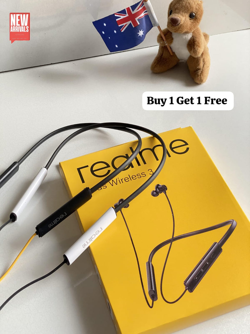 Realme wireless earbuds with a neckband-style cable and inline remote, displayed on top of a yellow Realme box. A small plush bear holding an Australian flag sits in the background on a white surface, with a 'Buy 1 Get 1 Free' label overlay.