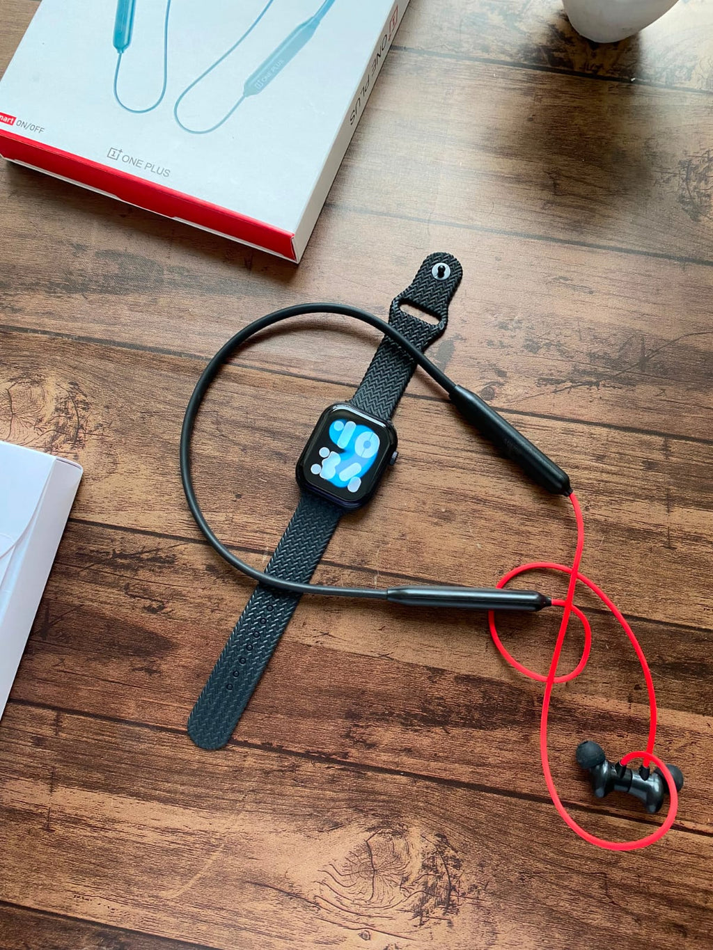 Black neckband-style smartwatch with a black woven strap and matching black wired earbuds, placed on a wooden tabletop. A white product box is partially visible in the background.