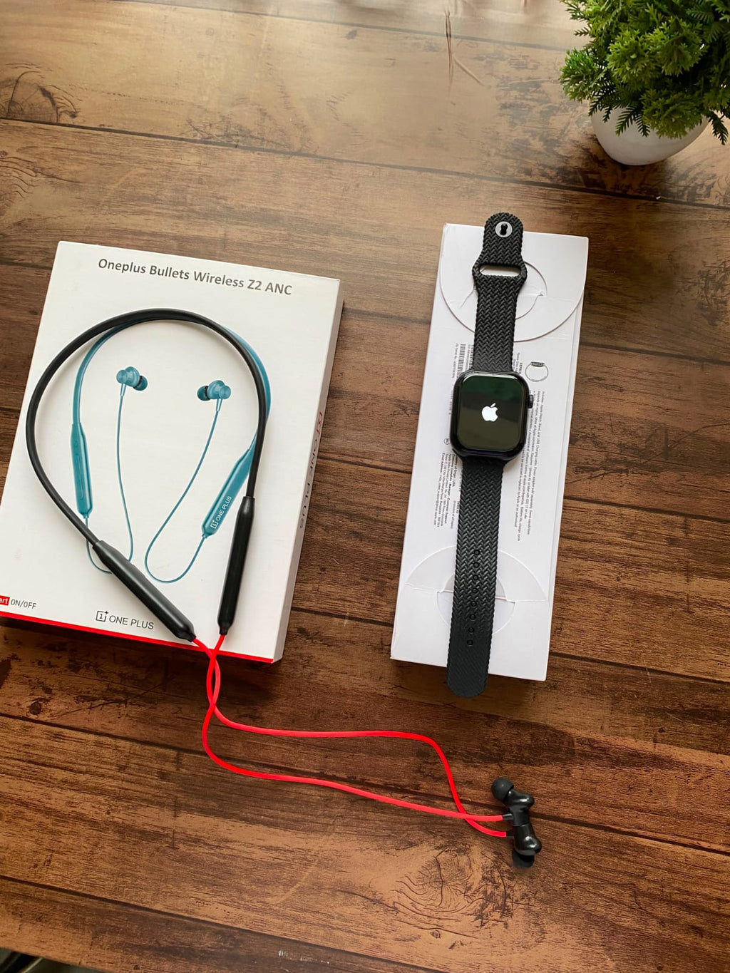 A pair of Oneplus Bullets Wireless 22 ANC in-tail earbuds with a black neckband and red cable, shown next to their white retail box on a wooden table. A black smartwatch is also displayed on a white box in the same setup.