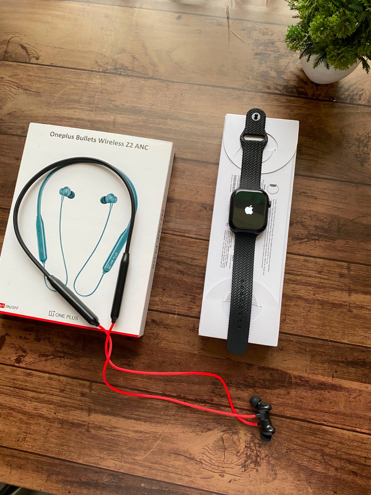 A pair of Oneplus Bullets Wireless 22 ANC in-tail earbuds with a black neckband and red cable, shown next to their white retail box on a wooden table. A black smartwatch is also displayed on a white box in the same setup.