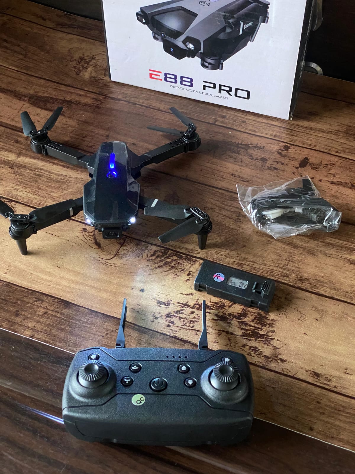 A black quadcopter drone with a blue LED light on top, shown with its remote controller, a small battery pack, and the product box on a wooden table.
