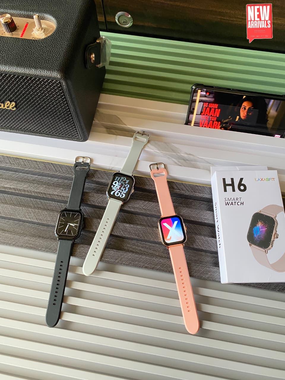 Three smartwatches with black, white, and peach straps are displayed on a striped surface next to their packaging box, with a speaker and a phone in the background.
