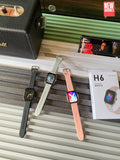 Three smartwatches with black, white, and peach straps are displayed on a striped surface next to their packaging box, with a speaker and a phone in the background.