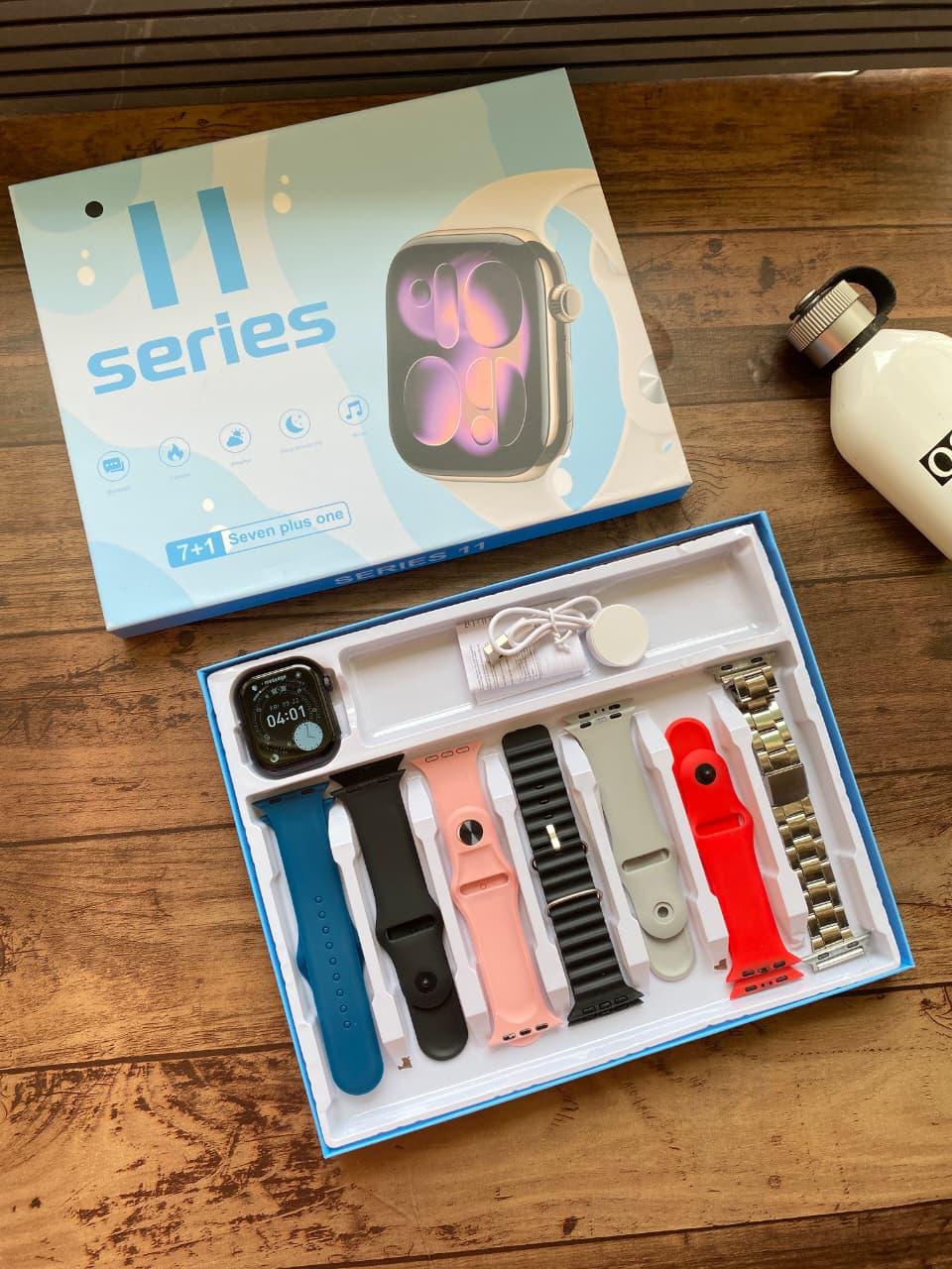 Apple Watch 11 Series smartwatch kit displayed on a wooden surface, showing the retail box, the open tray with a watch and multiple colorful bands, and a white bottle nearby.