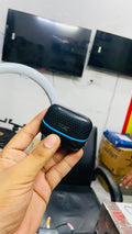 A hand holding a small black textured wireless earbud charging case with a bright blue accent line, set against a cluttered indoor background with computer monitors and office items.
