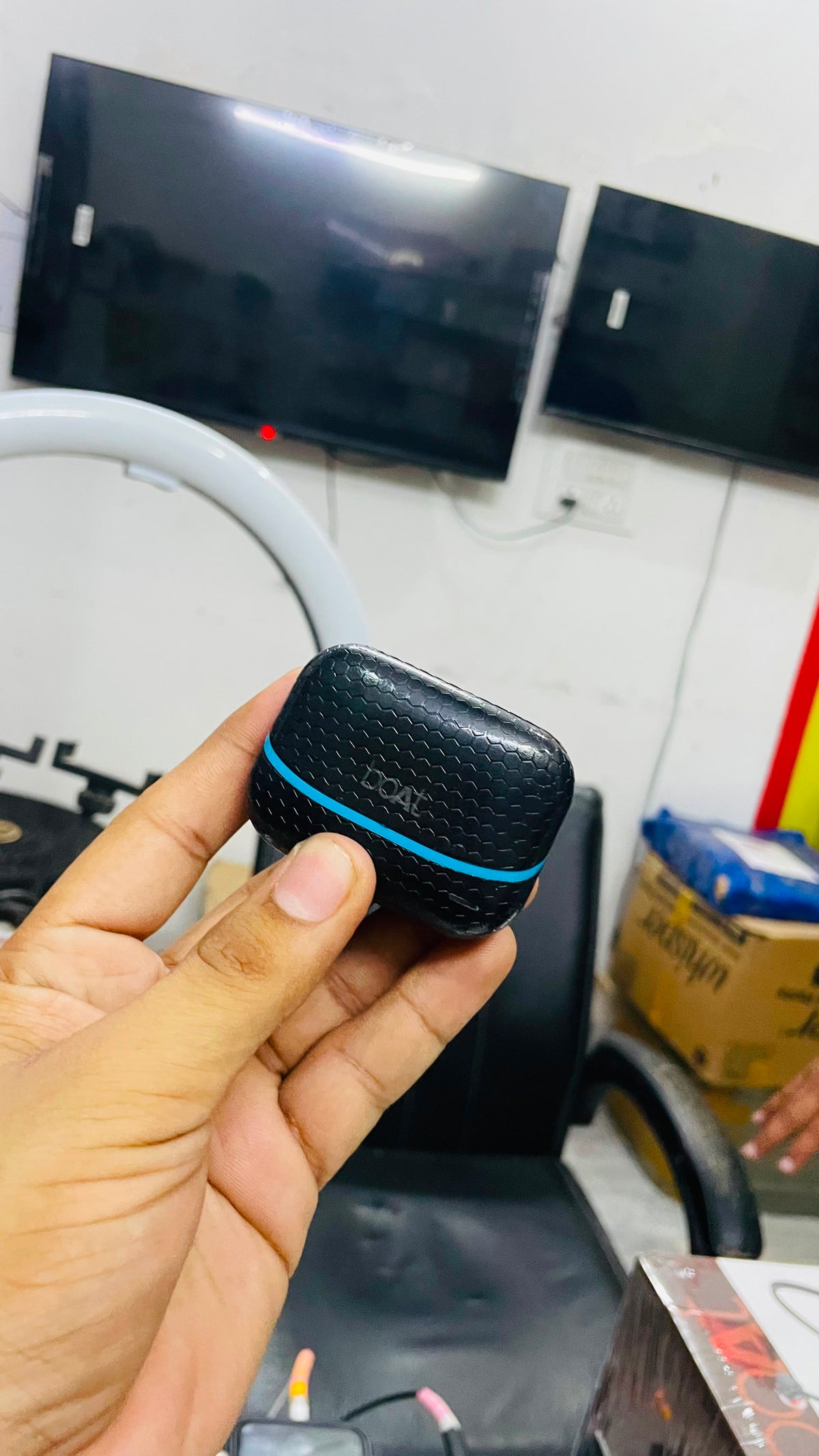 A hand holding a small black textured wireless earbud charging case with a bright blue accent line, set against a cluttered indoor background with computer monitors and office items.