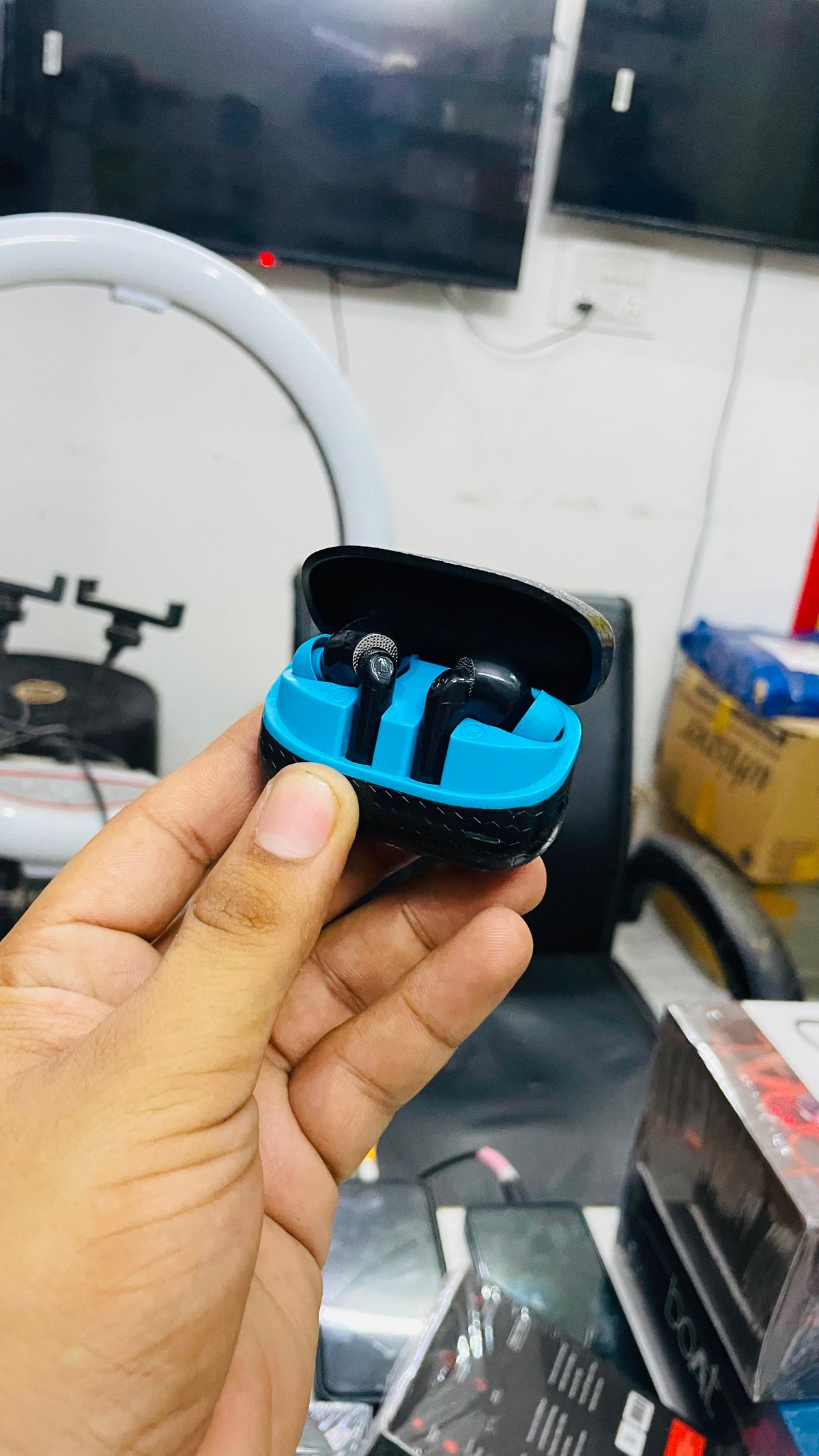 A hand holding a pair of black wireless earbuds inside an open black and bright blue charging case, with a retail store background visible.