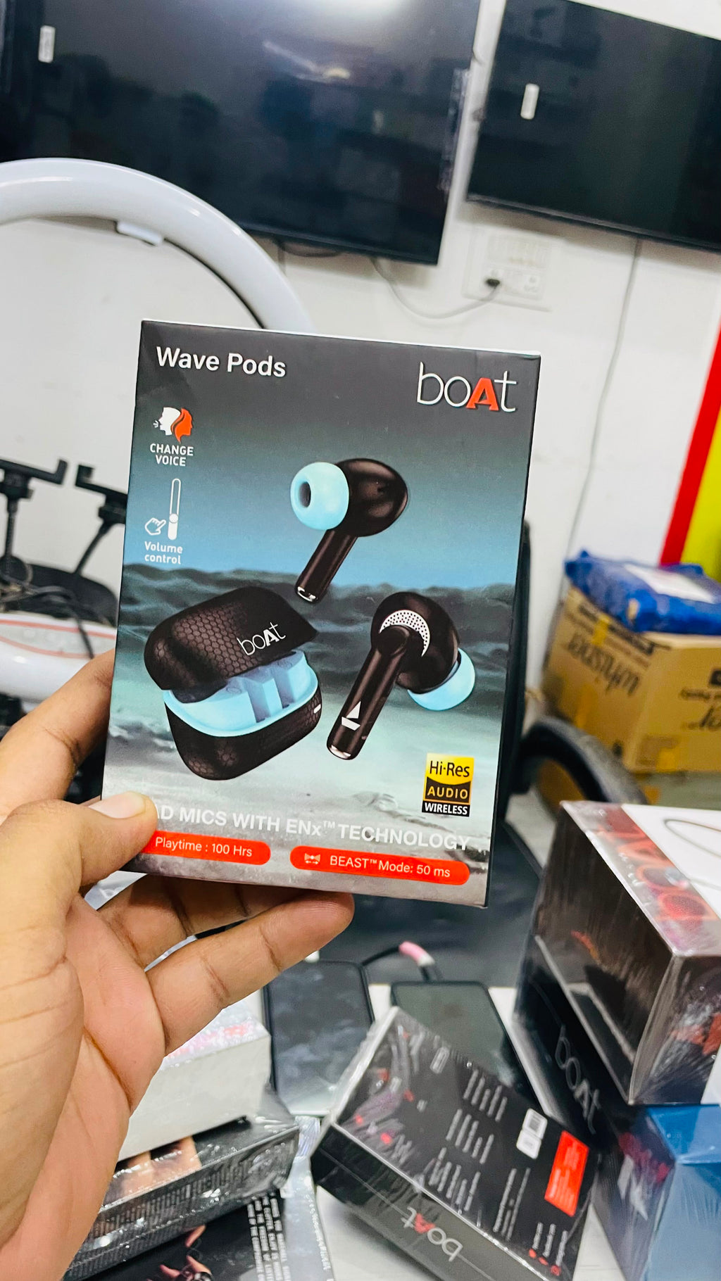 A hand holds a retail box of Boat Wave Pods wireless earbuds in a cluttered warehouse or showroom setting with other boxes and electronic equipment in the background.